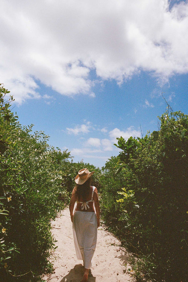 A woman walking on a sandy path wearing The Desert Cowboy straw fedora hat in natural color, surrounded by greenery and blue sky.