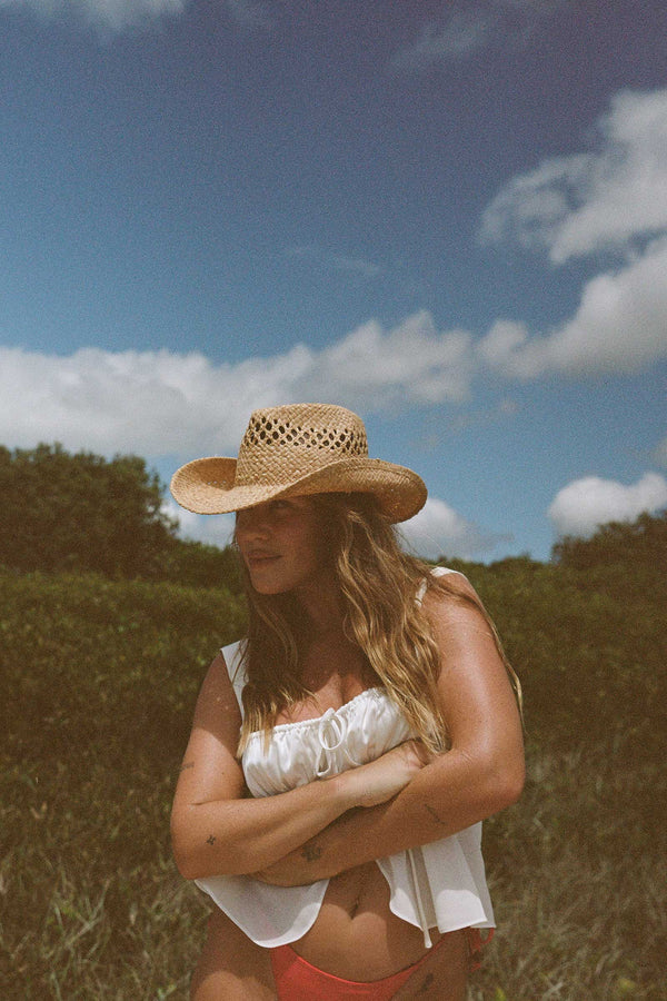 A woman wearing The Desert Cowboy straw fedora hat in natural color, standing outdoors with a green background.