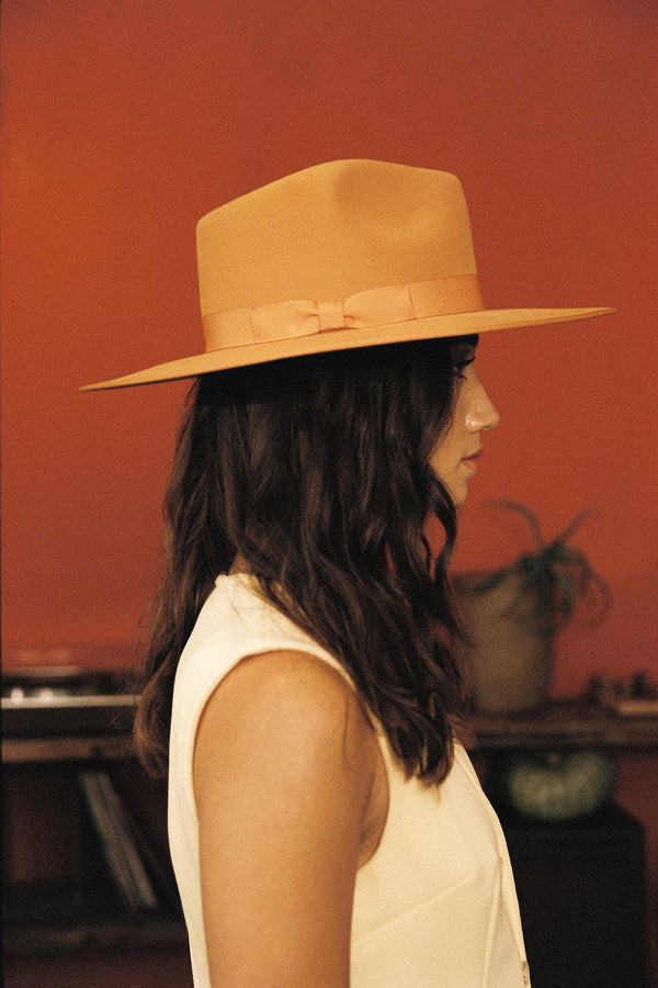 Desert Sun Rancher hat in brown, featuring a wide brim and tonal ribbon trim, worn by a model against an orange background.