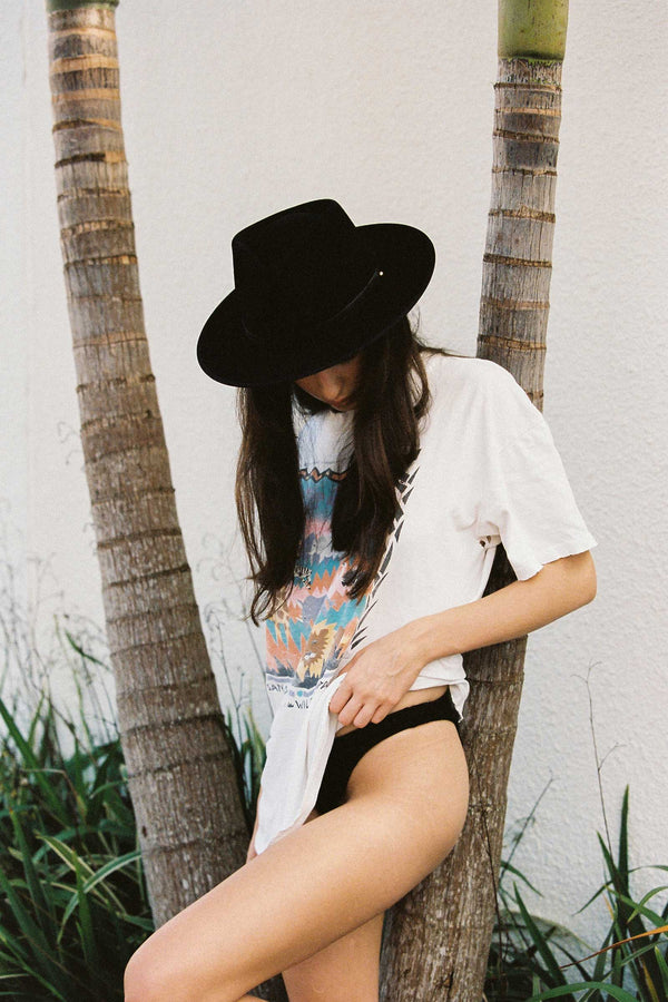 A woman wearing a black wool felt fedora hat, standing by a tree, with a graphic t-shirt and shorts.