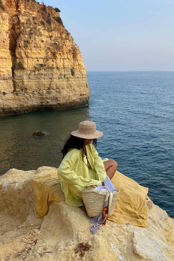 A woman sitting on a cliff by the sea wearing a natural straw Inca Bucket Wide hat, with a bag beside her.