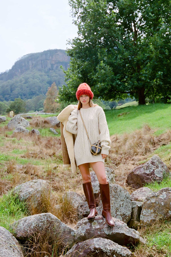 Model wearing a red Kai Beanie in a natural outdoor setting with a cream sweater and brown boots.