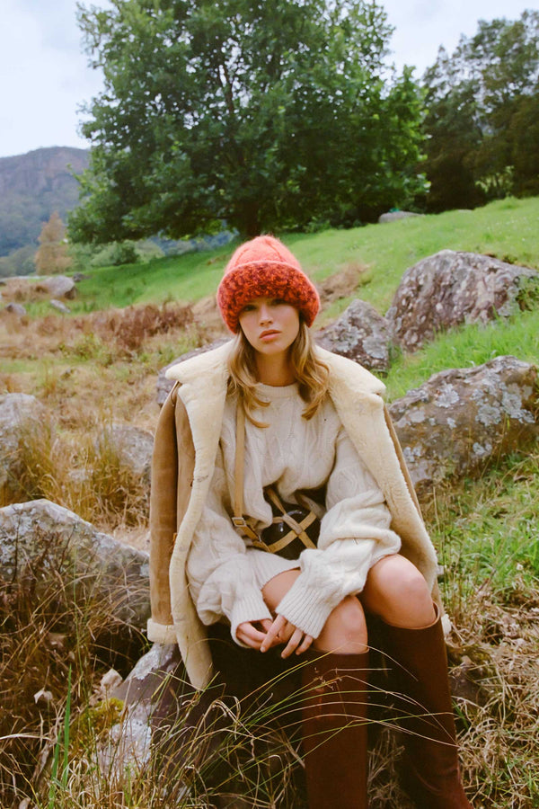 Model wearing a Kai Beanie in Flame color, sitting outdoors in a natural setting.