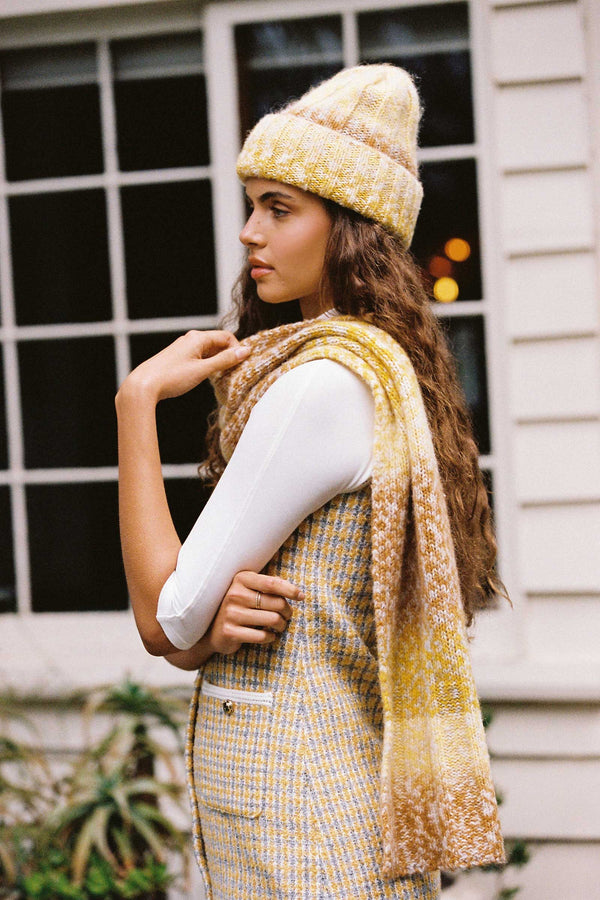 Model wearing a mustard and caramel ribbed scarf with a matching beanie, standing by a window.