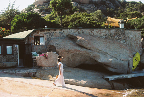 A person in a white dress and hat walking near a stone structure in a natural setting.