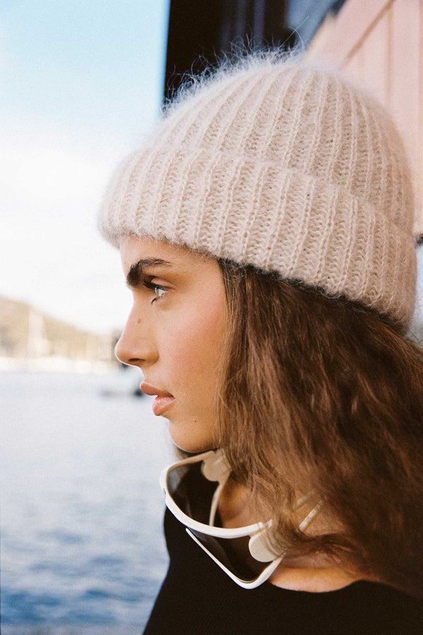 Profile view of a model wearing a natural-colored Mason Beanie by Lack of Color, with a scenic background.