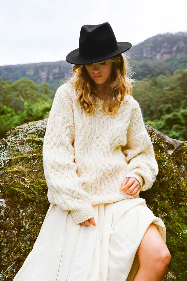 Model wearing a black wool felt Palo Fedora hat with a cream sweater and skirt, outdoors.