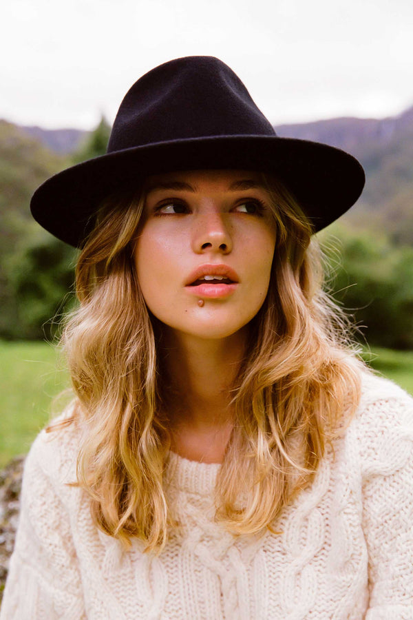 A woman wearing a black wool felt fedora hat, with wavy hair and a cream sweater, outdoors.