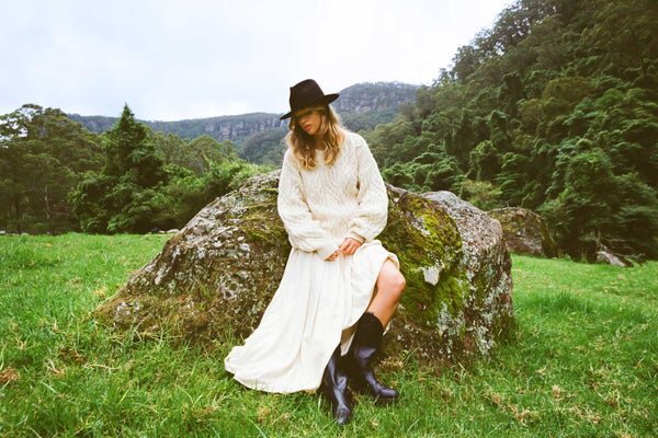 Model wearing The Palo Fedora in a natural setting, sitting on a rock, wearing a cream sweater and white skirt.