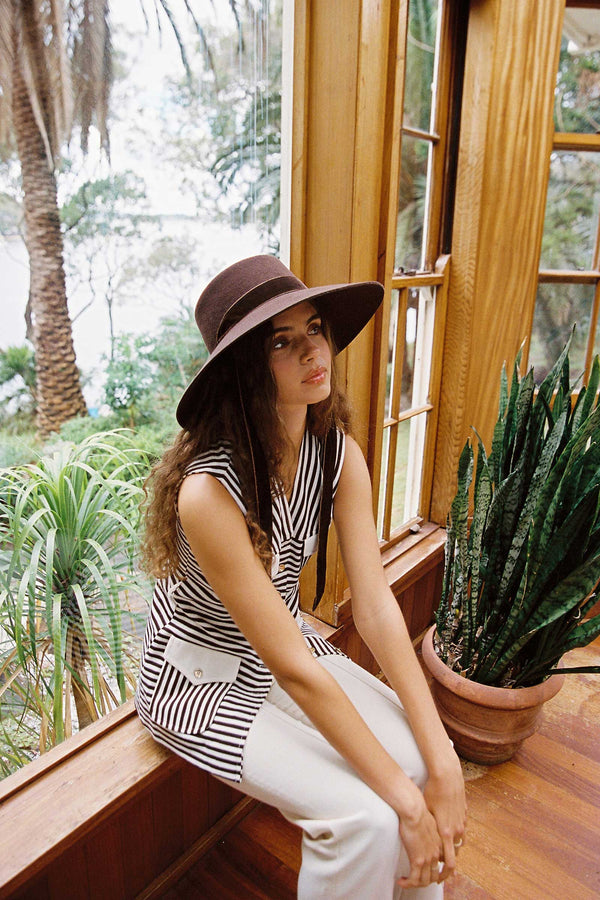 Model wearing the Paloma Midi Wool Felt Boater Hat in Brown, sitting in a sunlit room with plants.