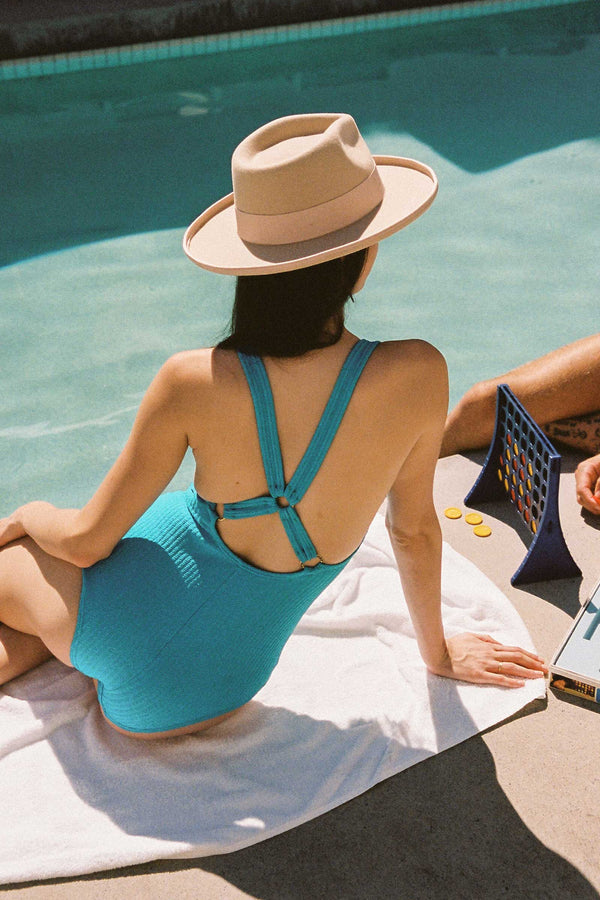 Woman wearing a pink fedora hat by the poolside, sitting on a towel.