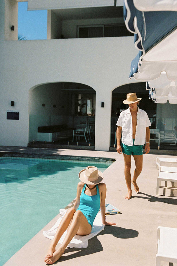 A woman in a blue swimsuit and a man in a white shirt and green shorts by a pool, both wearing beige fedora hats.