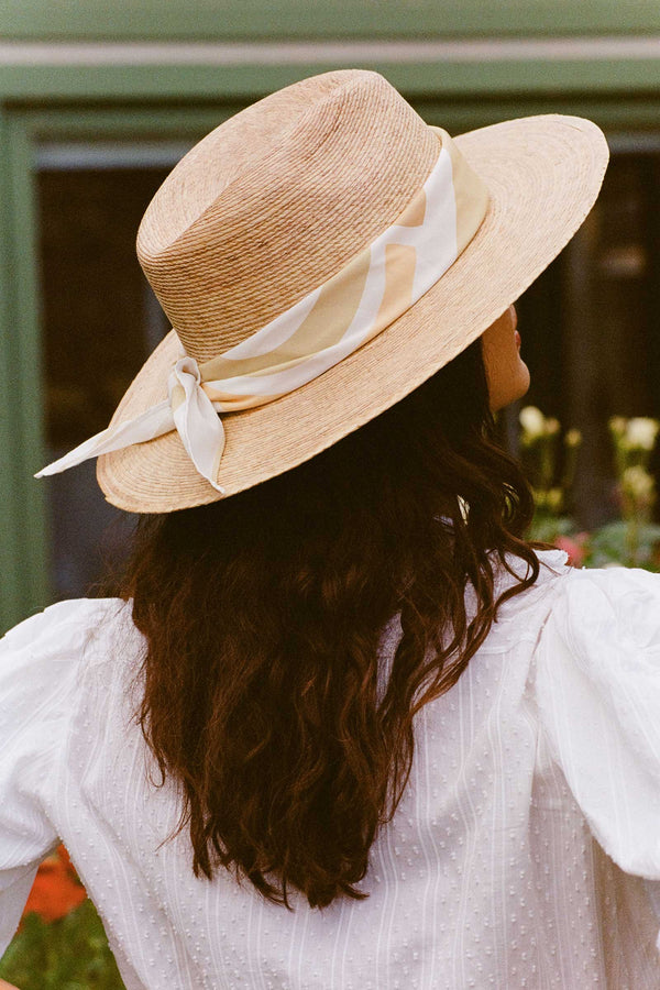 Woman wearing a straw hat with a cream and green scarf, viewed from behind.