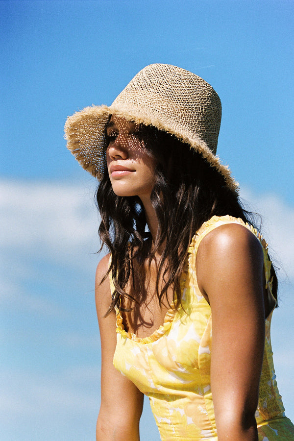 Model wearing a natural straw Reef Bucket hat against a blue sky, showcasing its frayed edge and casual style.
