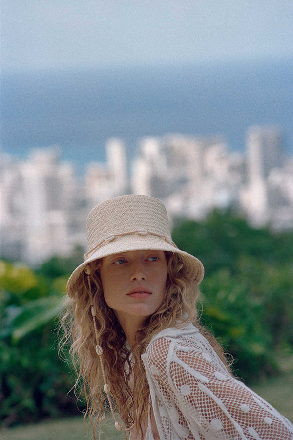 Model wearing The Inca Bucket straw hat with seashell chin strap against a city backdrop