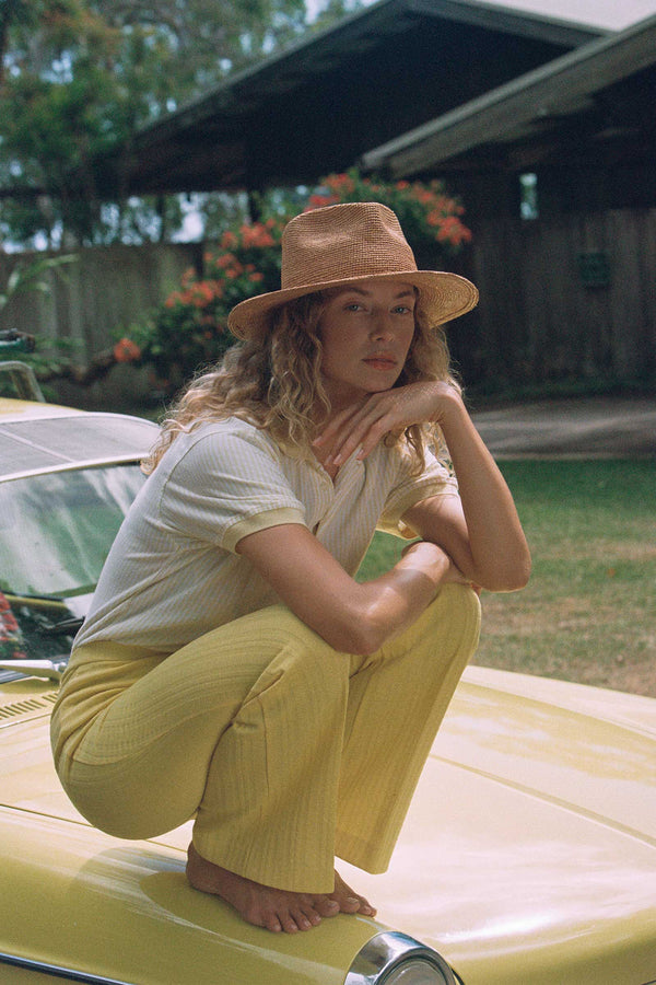 Model wearing The Inca Fedora, a natural straw hat, sitting on a yellow car in a tropical setting.