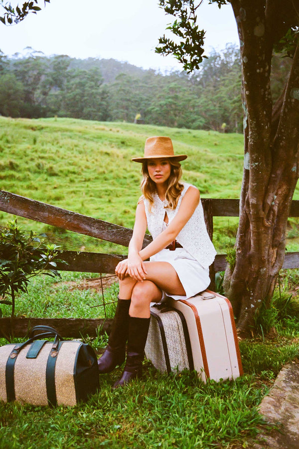 A model wearing The Inca Fedora, sitting on a suitcase in a green field.