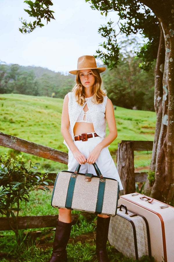 A model wearing The Inca Fedora, holding a suitcase in a green landscape.