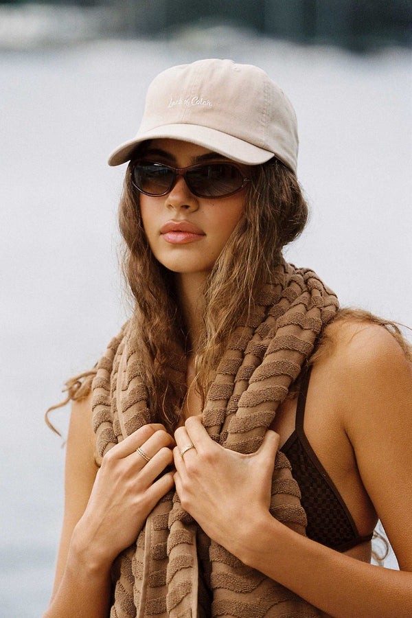 A model wearing a cream LOC Cap and sunglasses, holding a brown scarf by the water.
