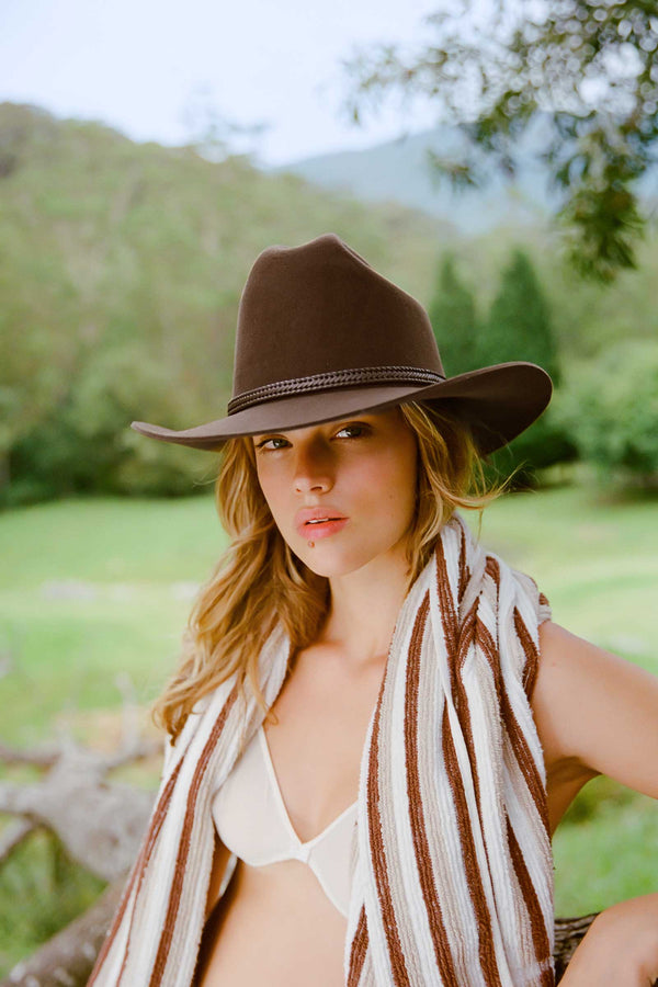 A woman wearing The Outback wool felt cowboy hat in brown, with a striped scarf, in a natural outdoor setting.