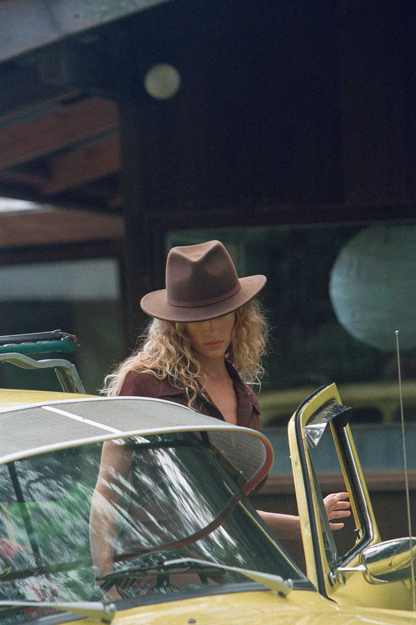 A model wearing a brown wool felt Palo Fedora hat, standing next to a yellow car.
