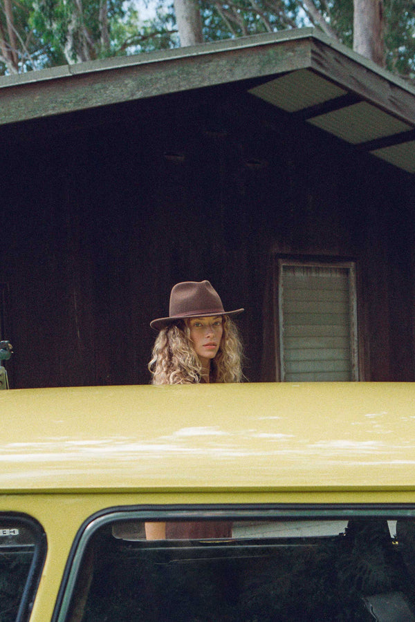 A woman with curly hair wearing a brown wool felt fedora, standing behind a yellow car near a wooden cabin.
