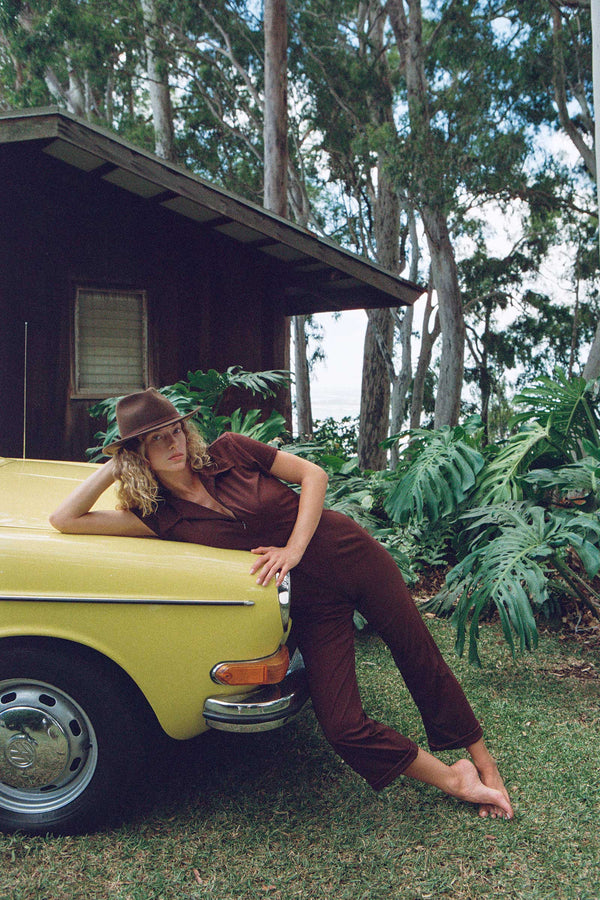 Model wearing the Palo Fedora in brown, leaning on a yellow car in a lush outdoor setting.