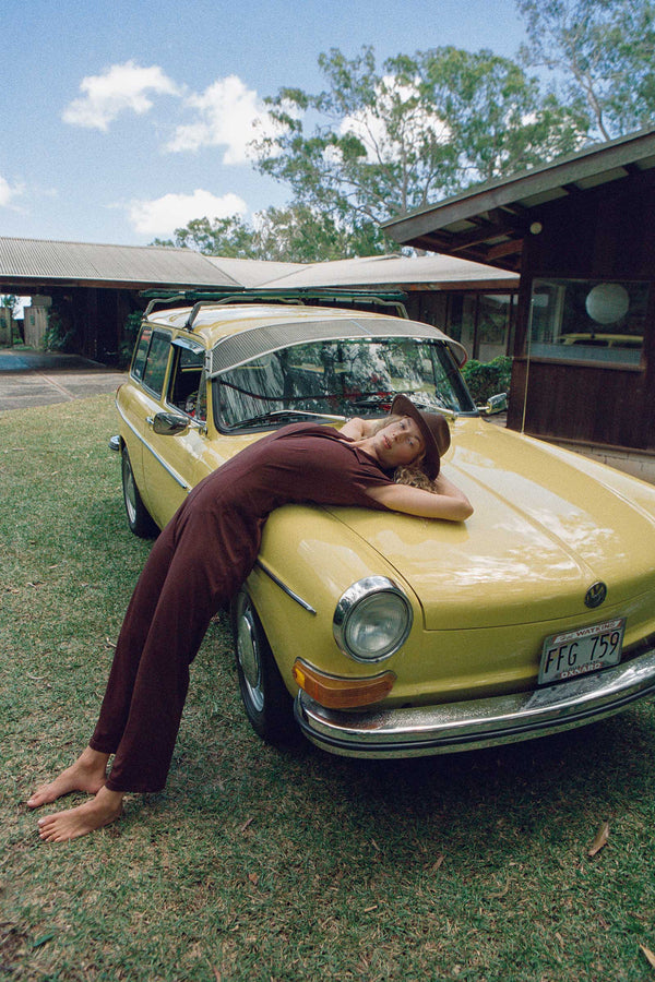 Model wearing the Palo Fedora, a brown wool felt hat, lounging on a yellow car in a natural setting.