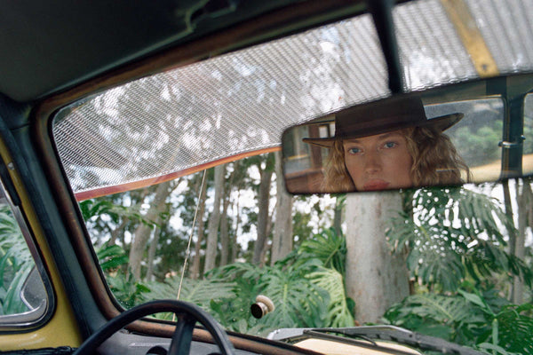 A woman wearing a brown Palo Fedora, seen in a car's rearview mirror with greenery in the background.