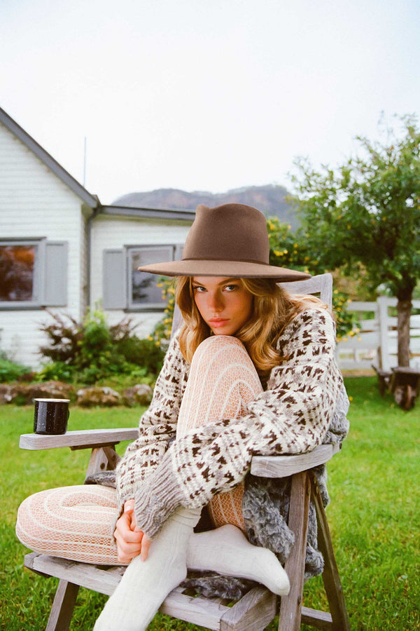 Model wearing The Weekender wool felt fedora hat in brown, sitting on a chair outdoors.