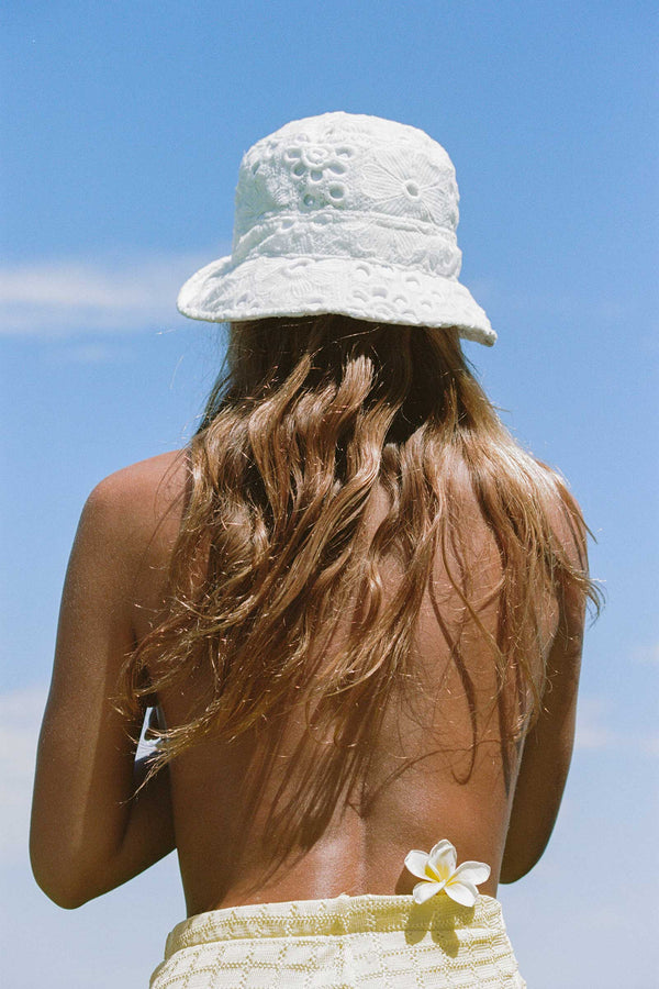 A woman with long hair wearing a white lace bucket hat, standing against a blue sky.