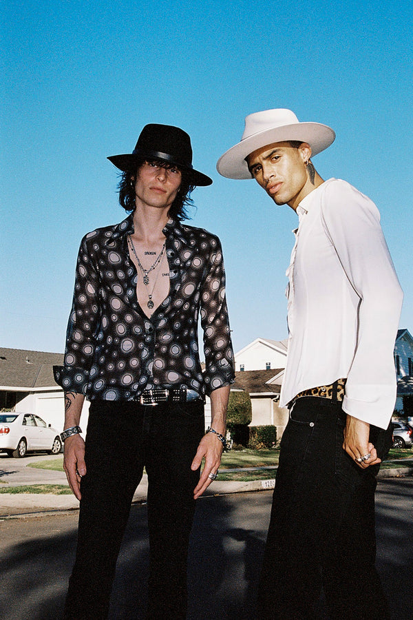 Two individuals wearing hats, one in a black fedora and the other in a white fedora, standing outdoors.