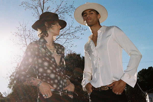 Two models wearing hats, one in a white fedora and the other in a brown hat, outdoors with sunlight.