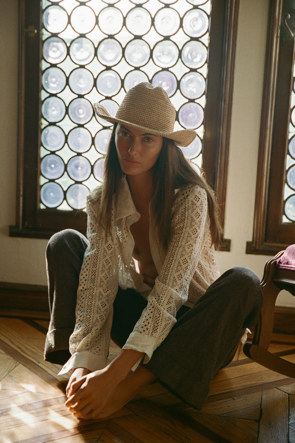 Model wearing a Raffia Cowboy straw hat, sitting on the floor in a sunlit room with patterned windows.