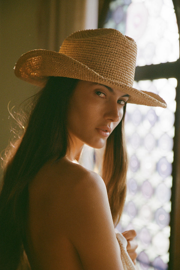 Model wearing a natural raffia cowboy hat, with a soft background and light filtering through a window.