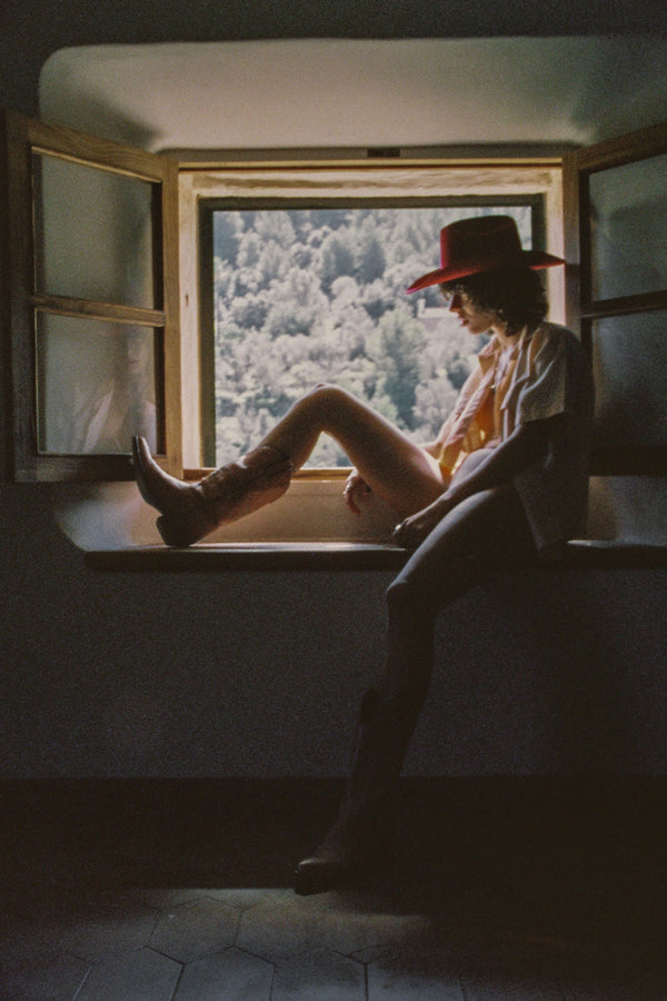 Model sitting by a window wearing a red wool felt cowboy hat and cowboy boots.
