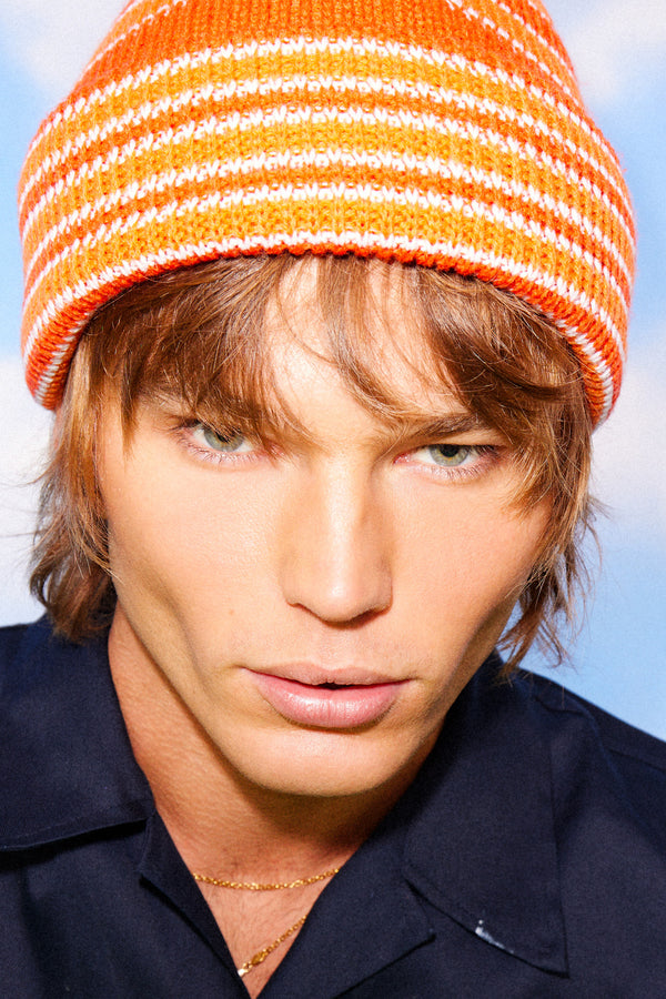 Model wearing Momo Beanie in orange stripes against a blue sky background