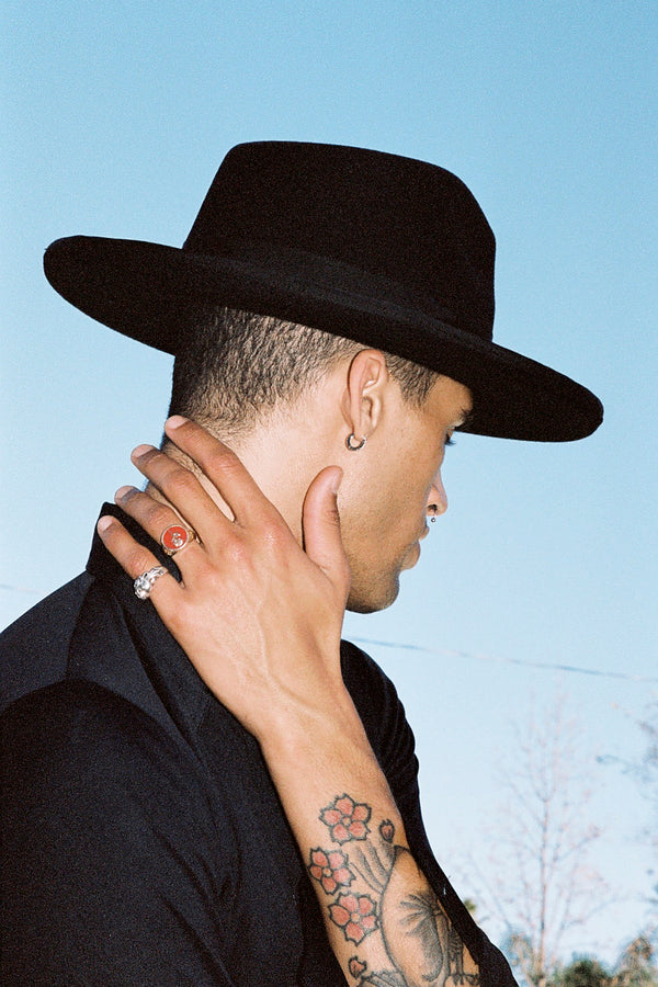 Model wearing a black wool felt fedora hat, showcasing a side profile with tattoos visible on the neck and hand.