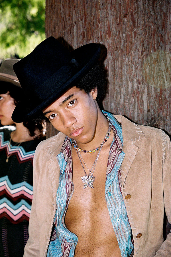 Model wearing a black wool felt fedora hat, styled with a colorful shirt and necklace, against a tree background.