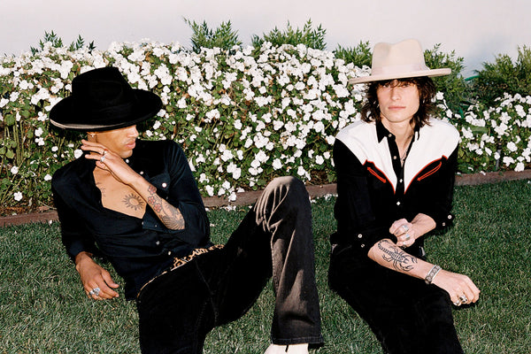 Two individuals wearing hats, one in a black fedora and the other in a beige hat, sitting on grass with flowers in the background.