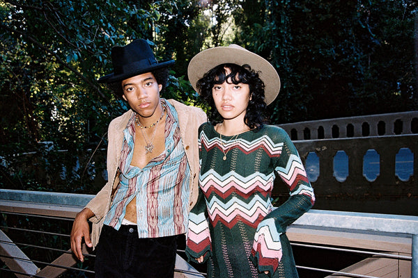 Two models wearing hats, one in a black fedora and the other in a beige hat, standing outdoors.