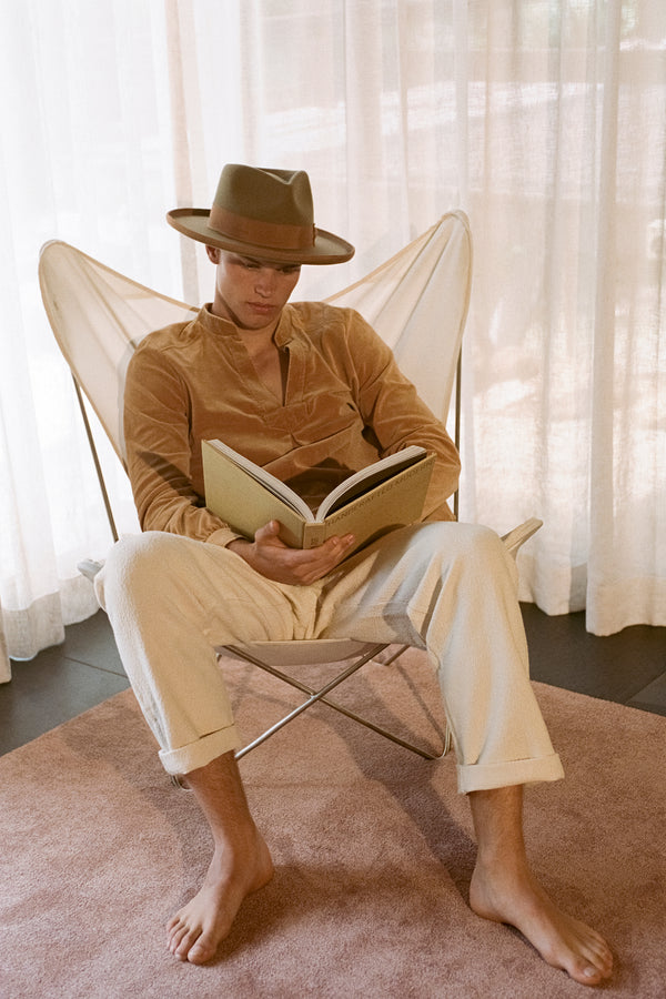 A man sitting in a chair wearing a brown fedora hat, reading a book.