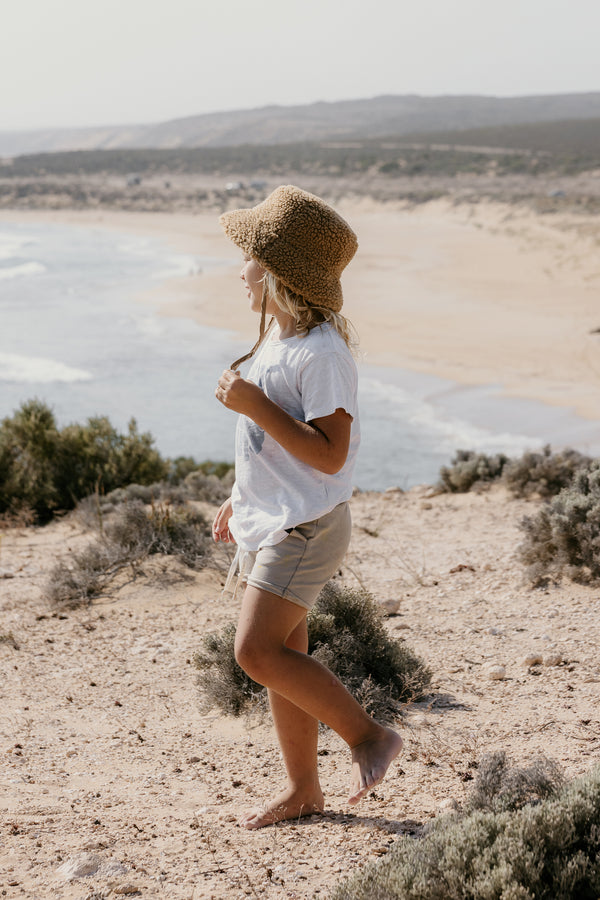 Child walking on a beach wearing a brown teddy bucket hat