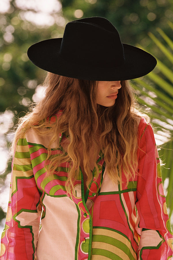 A woman wearing The Melodic Fedora in black, with a colorful shirt, in a natural setting.