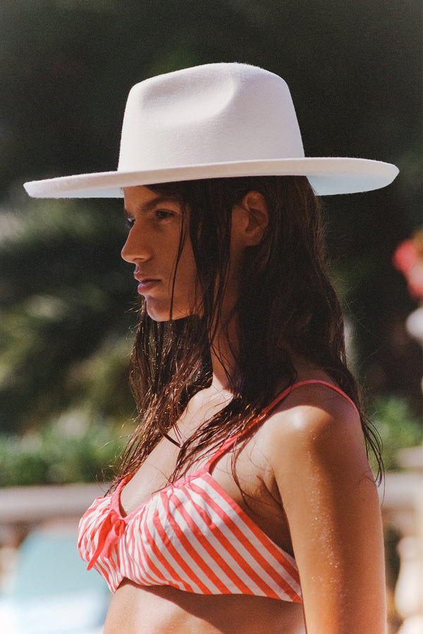 Model wearing The Melodic Fedora in white, with a striped bikini, outdoors.