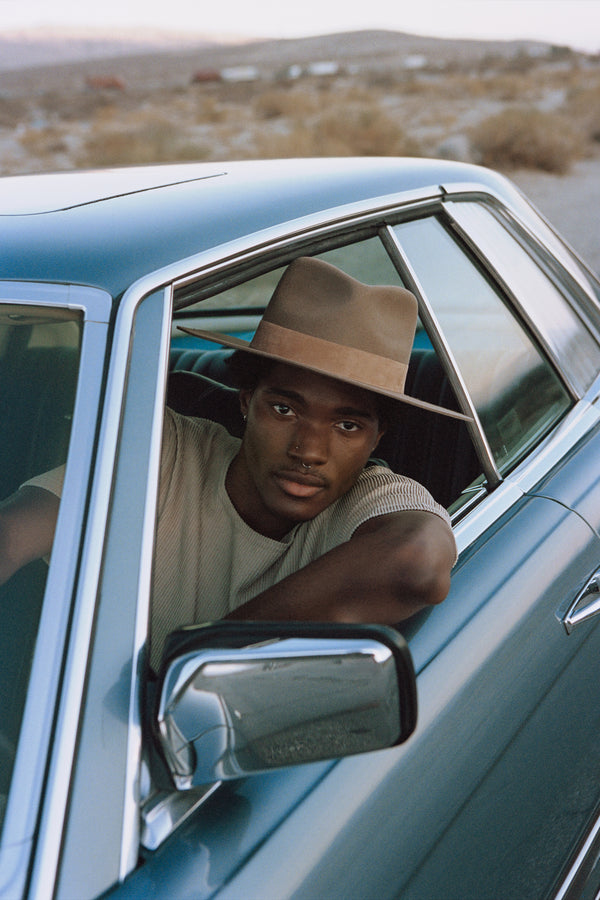 A model wearing The Mirage - Teak fedora hat while seated in a car.