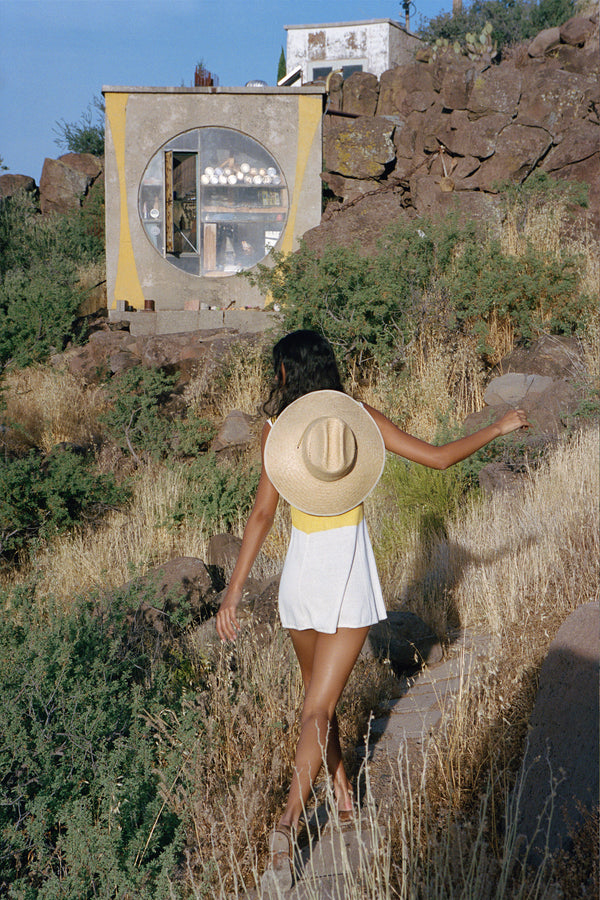 A woman walking outdoors wearing a wide-brimmed straw cowboy hat, with a natural landscape in the background.