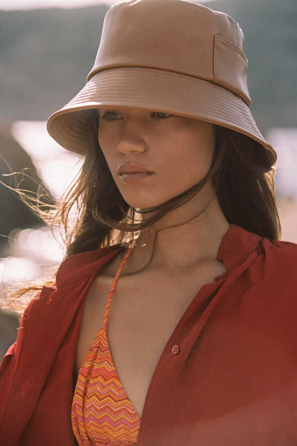 Model wearing a tan vegan leather bucket hat with a red shirt and a patterned bikini top, outdoors.
