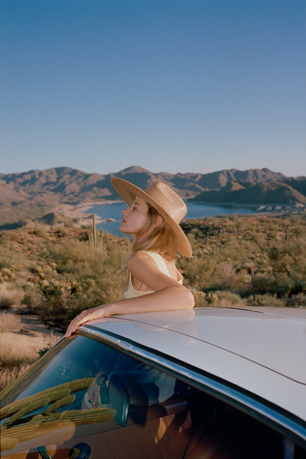 A woman wearing a straw cowboy hat, leaning out of a car in a desert landscape with mountains in the background.