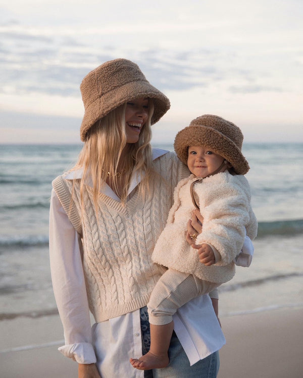 A woman and a child wearing brown teddy bucket hats on the beach at sunset.